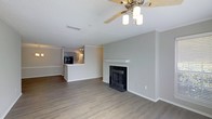 This single-story space showcases an open-concept layout that seamlessly connects the living area to the kitchen. The living room serves as the focal point of the home, highlighted by a classic fireplace with a white painted mantel and dark surround, providing both a functional and aesthetic centerpiece. A ceiling fan with integrated lighting is mounted overhead, ensuring year-round comfort and ambient illumination throughout the space.

The flooring throughout the living area consists of dark hardwood-style planks, offering a warm yet contemporary feel that contrasts cleanly with the light grey walls and crisp white baseboards and crown molding. A textured ceiling adds subtle visual interest above.

The kitchen is accessible via a raised pass-through counter, which defines the boundary between the two spaces while maintaining an open, connected feel. The kitchen is equipped with dark cabinetry and black appliances, visible from the living area, and is finished with track lighting overhead. The overall color palette — neutral greys, white trim, and rich dark flooring — gives the space a modern, cohesive aesthetic that is both practical and visually appealing.
