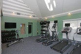 This fitness center offers a thoughtfully designed workout environment finished with soft mint-green walls and neutral carpeted flooring that creates a clean, energizing atmosphere. Track lighting along the ceiling provides ample illumination throughout the space, complemented by natural light filtering in through large windows dressed with roll-down shades. The room is equipped with a selection of cardio machines, including elliptical trainers and a treadmill, each outfitted with individual display monitors for an enhanced workout experience. A dedicated strength and functional training area features a multi-tiered equipment rack stocked with medicine balls and kettlebells of varying weights, alongside a TRX suspension training system mounted to the wall. A wall-mounted exercise mat is also available for stretching or floor-based routines. The layout is open and well-organized, allowing users to move freely between equipment. Tray ceiling detailing adds a subtle architectural element, lending the room a polished, finished look. Overall, the space balances practicality with comfort, making it suitable for a range of fitness routines from cardio and strength training to flexibility work.