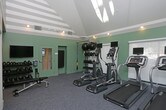 This fitness center offers a thoughtfully designed workout environment finished with soft mint-green walls and neutral carpeted flooring that creates a clean, energizing atmosphere. Track lighting along the ceiling provides ample illumination throughout the space, complemented by natural light filtering in through large windows dressed with roll-down shades. The room is equipped with a selection of cardio machines, including elliptical trainers and a treadmill, each outfitted with individual display monitors for an enhanced workout experience. A dedicated strength and functional training area features a multi-tiered equipment rack stocked with medicine balls and kettlebells of varying weights, alongside a TRX suspension training system mounted to the wall. A wall-mounted exercise mat is also available for stretching or floor-based routines. The layout is open and well-organized, allowing users to move freely between equipment. Tray ceiling detailing adds a subtle architectural element, lending the room a polished, finished look. Overall, the space balances practicality with comfort, making it suitable for a range of fitness routines from cardio and strength training to flexibility work.
