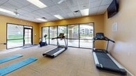 This fitness center is a comprehensively outfitted workout facility designed to support a wide range of exercise routines. The space features warm golden-yellow walls, a drop-tile ceiling with recessed fluorescent lighting, and low-pile carpet flooring throughout, creating a clean and energizing atmosphere.

The cardio zone is well-appointed with multiple treadmills, a stationary bike, an elliptical trainer, and a stair climber, all positioned to take advantage of the room's standout feature: a full wall of large floor-to-ceiling glass windows and sliding doors. These expansive windows flood the space with natural light and offer direct views of an outdoor pool and covered courtyard with arched columns and landscaping, creating an inviting and open feel during workouts.

A dedicated strength training area houses a multi-station cable machine with attachments for a variety of upper and lower body exercises, accompanied by an adjustable bench. A wall-mounted flat-screen television is positioned nearby for entertainment.

The functional fitness area is stocked with exercise mats, stability balls in multiple sizes, resistance bands, foam rollers, and a dedicated storage rack, making the space well-suited for stretching, core work, and flexibility training. A ceiling fan adds additional air circulation. Steps visible in the background suggest connectivity to an adjacent or elevated area within the facility.