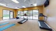 This fitness center is a comprehensively outfitted workout facility designed to support a wide range of exercise routines. The space features warm golden-yellow walls, a drop-tile ceiling with recessed fluorescent lighting, and low-pile carpet flooring throughout, creating a clean and energizing atmosphere.

The cardio zone is well-appointed with multiple treadmills, a stationary bike, an elliptical trainer, and a stair climber, all positioned to take advantage of the room's standout feature: a full wall of large floor-to-ceiling glass windows and sliding doors. These expansive windows flood the space with natural light and offer direct views of an outdoor pool and covered courtyard with arched columns and landscaping, creating an inviting and open feel during workouts.

A dedicated strength training area houses a multi-station cable machine with attachments for a variety of upper and lower body exercises, accompanied by an adjustable bench. A wall-mounted flat-screen television is positioned nearby for entertainment.

The functional fitness area is stocked with exercise mats, stability balls in multiple sizes, resistance bands, foam rollers, and a dedicated storage rack, making the space well-suited for stretching, core work, and flexibility training. A ceiling fan adds additional air circulation. Steps visible in the background suggest connectivity to an adjacent or elevated area within the facility.