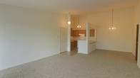 This single-floor residence offers a clean, open layout that connects the living area to the kitchen in a practical and functional design. The spacious living area is finished with neutral-toned carpet, providing a warm and comfortable foundation throughout the space. A smooth, light-colored ceiling carries two pendant light fixtures — a simple globe pendant and a small chandelier — that help delineate the living and kitchen zones while adding a touch of character overhead.

The kitchen is positioned at the rear of the open-plan space and features warm medium-toned wood cabinetry along the upper and lower walls, offering ample storage. A white appliance suite complements the light countertops, creating a bright and clean aesthetic. A peninsula with a raised breakfast bar separates the kitchen from the living area, serving as both a functional workspace and a casual dining or gathering spot.

The transition between the carpeted living area and the tile flooring of the kitchen is clearly defined, adding visual distinction between the two zones. A white paneled door provides access to an adjacent area, contributing to the overall flow of the layout. The neutral color palette throughout — whites, creams, and soft tones — gives the space a bright, airy ambiance with a timeless, understated appeal.