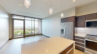 Located at 180 E Water Street in downtown Kalamazoo, Michigan, this contemporary single-floor unit offers a well-appointed layout across six distinct spaces. The open-concept kitchen is a standout feature, showcasing rich dark wood cabinetry that extends to the ceiling, quartz countertops, a geometric white tile backsplash, and a full suite of stainless steel appliances including a gas range, built-in microwave, and French door refrigerator. A central island with a light quartz surface provides additional prep and gathering space, illuminated by a trio of modern geometric pendant lights overhead. Light hardwood flooring runs throughout, complementing the neutral gray walls and dark baseboard trim for a cohesive, modern aesthetic. The living area is defined by expansive floor-to-ceiling windows that flood the space with natural light and offer elevated views of the Kalamazoo skyline and surrounding greenery, with a glass-railed balcony accessible via sliding doors. The bedroom features generous closet space and its own access to the curtain wall windows. The bathroom is finished with large-format tile flooring, a navy blue vanity with white countertop, and backlit mirrors framed by sleek wall sconces, lending a polished, hotel-inspired feel to the space.