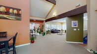 Located at 1055 S Zeno Way in Aurora, Colorado, this single-floor facility is defined by its striking architectural character and thoughtfully organized communal areas. The central gathering space commands attention with dramatically vaulted ceilings accented by bold dark wood beams, creating a sense of openness and grandeur. Warm terracotta and sage green accent walls lend the interior a welcoming, colorful energy throughout.

The main open area flows naturally into distinct functional zones. A lounge area features rich dark leather seating arranged around a coffee table, surrounded by large windows dressed with wood-toned blinds that fill the room with soft, diffused natural light and offer views of the surrounding exterior. Lush potted plants are distributed throughout, adding warmth and vitality to the space.

A dining area accommodates small groups with table-and-chair sets, while a compact, fully equipped kitchen includes wood cabinetry, tile flooring, a double-basin sink, and standard appliances — ideal for light food preparation. A quiet nook with wraparound windows and a classic wooden chair provides a cozy retreat for reading or reflection.

Additional areas visible in the layout suggest dedicated spaces for desk work or administrative use. Plush neutral carpeting runs throughout the main areas, tying together the varied zones into a cohesive, functional, and comfortable environment.