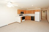 This single-floor unit offers a straightforward, functional layout designed for comfortable everyday living. The living area is generously proportioned, featuring neutral-toned carpet, a ceiling fan with integrated lighting, a textured ceiling, and a front entry door with a transom window that allows natural light to filter in. A window with blinds and a wall-mounted air conditioning unit provide climate control options alongside the baseboard heating system found throughout the unit.

The kitchen is well-equipped and efficiently arranged in an L-shaped configuration. Warm oak cabinetry extends to the upper and lower walls, offering substantial storage space. Light-toned countertops provide ample workspace, and the kitchen includes a double-basin stainless steel sink, an electric range, a built-in over-the-range microwave, and a black dishwasher. The kitchen opens directly to the main living space, creating an connected feel between the two areas.

The bathroom is clean and practical, featuring light tile flooring, a wide vanity with a white countertop and oak cabinetry below, a large mirror, and a full-size tub and shower combination. The overall aesthetic throughout the unit is neutral and understated, with warm wood tones providing visual consistency.