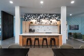 This single-floor communal space is designed with a contemporary open-plan layout that seamlessly integrates a fully equipped kitchen, multiple dining areas, and a comfortable lounge. Polished concrete floors extend throughout, reflecting natural light and reinforcing the space's clean, modern aesthetic.

The kitchen is a standout feature, anchored by a large white quartz island with bar seating. Light wood cabinetry, stainless steel appliances, and an eye-catching geometric hexagonal tile backsplash that extends dramatically to the ceiling give the kitchen a bold, artistic character. Wall-mounted sconce lighting adds warmth to the space.

The dining area accommodates multiple large tables with modern seating, illuminated by circular ring pendant lights suspended from the ceiling. Large windows with roller shades line the walls, providing generous natural light and street-level views of the surrounding neighbourhood.

The lounge area features a grey sectional sofa, a wall-mounted flat-screen television set against a teal geometric accent wall, and a pair of bold blue accent chairs positioned near floor-to-ceiling windows. Recycling and waste stations are conveniently integrated near the dining area. The overall ambiance is bright, functional, and thoughtfully designed for community use.