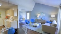 This single-floor space makes a striking first impression with its bold, cohesive design aesthetic. The living area is the centerpiece, featuring rich hardwood flooring, vaulted ceilings that add a sense of openness, and walls painted in a calming slate blue that ties the entire space together. Vivid cobalt blue accent chairs paired with a geometric patterned area rug bring energy and personality to the room, while a low-profile coffee table and a warm wood-toned dresser provide both function and style. Curated wall art in complementary blues and greens adds an artistic touch throughout.

Natural light flows in through large windows dressed with flowing curtains, brightening the interior and enhancing the airy feel of the space. A dedicated nook area, visible from the main living space, features a desk and seating — ideal for a home office or study corner. The open layout seamlessly connects the living area to the kitchen, which is outfitted with light wood cabinetry and a clean countertop workspace. Yellow accent pieces scattered throughout add cheerful contrast to the dominant cool-toned palette, creating a lively yet balanced atmosphere that feels both modern and welcoming.