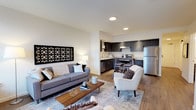 This single-floor space offers a thoughtfully designed open-concept layout that seamlessly connects the living area and kitchen, creating a bright and airy atmosphere ideal for both relaxing and entertaining. The living area is anchored by a sleek grey sofa and a complementary accent chair, arranged around a mid-century modern wood coffee table atop a patterned area rug. A striking geometric wall art piece adds a bold focal point to the neutral, light-toned walls, while recessed lighting overhead ensures a well-lit and inviting ambiance. Warm hardwood-style flooring flows throughout, adding continuity and a sense of warmth to the space. The kitchen is outfitted with dark cabinetry, stainless steel appliances including a full-size refrigerator, and a functional island with bar seating — making it both practical and stylish. Clean white countertops contrast beautifully against the dark cabinetry, lending a contemporary aesthetic. The open sightlines between the kitchen and living area enhance the overall sense of spaciousness, making this well-appointed unit feel larger than its footprint suggests. The overall design balances modern minimalism with functional comfort.