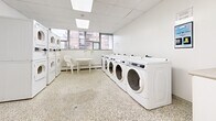 Located at 30 Queen St E in Mississauga, Ontario, this communal laundry facility offers a clean, well-organized space designed to serve building residents efficiently. The room features durable terrazzo flooring throughout, providing a practical and easy-to-maintain surface. The drop ceiling with integrated skylight panels allows natural light to filter in, brightening the white-painted interior and contributing to a clean, functional atmosphere.

The laundry area is equipped with a row of full-size front-loading washing machines along one wall, complemented by a set of stacked washer-dryer units, offering ample capacity for residents. A utility sink with cabinetry provides additional functionality for hand-washing or other laundry tasks. A fire extinguisher is prominently mounted near the entry door, ensuring safety compliance.

A row of windows along one wall frames views of the surrounding neighbourhood, allowing natural light to fill the space and creating an open, airy feel. Beneath the windows, baseboard heating ensures year-round comfort. A small round table with chairs provides a convenient waiting area for residents. The room is accessed through a solid door with a window panel, and an additional interior door leads to a secondary area. Floor drains are integrated into the terrazzo floor, reflecting the practical, utility-focused design of this well-maintained shared amenity space.