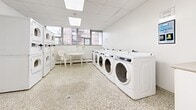 Located at 30 Queen St E in Mississauga, Ontario, this communal laundry facility offers a clean, well-organized space designed to serve building residents efficiently. The room features durable terrazzo flooring throughout, providing a practical and easy-to-maintain surface. The drop ceiling with integrated skylight panels allows natural light to filter in, brightening the white-painted interior and contributing to a clean, functional atmosphere.

The laundry area is equipped with a row of full-size front-loading washing machines along one wall, complemented by a set of stacked washer-dryer units, offering ample capacity for residents. A utility sink with cabinetry provides additional functionality for hand-washing or other laundry tasks. A fire extinguisher is prominently mounted near the entry door, ensuring safety compliance.

A row of windows along one wall frames views of the surrounding neighbourhood, allowing natural light to fill the space and creating an open, airy feel. Beneath the windows, baseboard heating ensures year-round comfort. A small round table with chairs provides a convenient waiting area for residents. The room is accessed through a solid door with a window panel, and an additional interior door leads to a secondary area. Floor drains are integrated into the terrazzo floor, reflecting the practical, utility-focused design of this well-maintained shared amenity space.