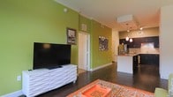 This single-floor residence makes a striking first impression with its bold, chartreuse green accent walls that energize the open-concept living space. Dark hardwood flooring runs throughout, providing a rich contrast to the bright walls and crisp white trim and crown molding that frame each room with a polished finish.

The living area is generously proportioned, offering ample space for comfortable furnishings. A sleek white media console anchors one wall, complemented by decorative wall art that adds personality and character to the space. The layout flows naturally toward the kitchen, which is visible from the living area and features dark cabinetry, stainless steel appliances including a refrigerator, and a breakfast bar with pendant lighting overhead. The warm glow of the hanging pendant lights adds an inviting ambiance to the kitchen zone.

The open sightlines between the living and kitchen areas create a connected, social atmosphere well-suited for both everyday living and entertaining. Interior doors with classic white paneling provide access to additional rooms, maintaining the clean and cohesive aesthetic throughout. The combination of vibrant color choices, contemporary finishes, and functional layout makes this a dynamic and visually engaging living space.
