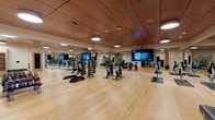 This well-appointed fitness studio is designed to accommodate a wide range of workout activities within a generously proportioned, open-plan layout. The space is immediately distinguished by its warm, wood-paneled ceiling, which adds a refined, contemporary aesthetic and contributes to the room's inviting atmosphere. Smooth hardwood floors run throughout, providing an ideal surface for both equipment-based and free-form exercise.

Full-length mirrors line the walls, visually expanding the space while allowing users to monitor their form during workouts. The studio is equipped with multiple spin bikes arranged for group cycling classes, along with a tiered dumbbell rack offering a comprehensive range of free weights. Large flat-screen displays are mounted throughout the room, capable of streaming workout classes or fitness content.

A dedicated storage area along one wall features built-in open shelving for organizing accessories, accompanied by a padded bench for convenience. Nearby, a well-stocked equipment rack holds stability balls, resistance bands, medicine balls, weighted sandbags, and step platforms, supporting a diverse range of training modalities.

Recessed drum pendant lighting provides consistent, even illumination throughout the studio. The overall design balances functionality with a polished, upscale finish, creating an environment well-suited for both individual training and group fitness sessions.