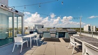 Located atop a modern multi-story building at 1970 N Milwaukee Ave in Chicago's vibrant Bucktown/Logan Square neighborhood, this rooftop deck offers an expansive outdoor amenity space with impressive, unobstructed views of the Chicago skyline and surrounding cityscape. The deck is finished with large-format concrete pavers, providing a clean, contemporary aesthetic that complements the building's modern architectural design.

The outdoor space is equipped with multiple grilling stations featuring built-in gas grills and tiled countertops, making it well-suited for outdoor cooking and entertaining. White modern dining tables and chairs are arranged throughout the deck, offering comfortable seating areas beneath overhead string lights that add a welcoming ambiance during evening gatherings. Planters with greenery are thoughtfully integrated into the space, adding natural elements to the rooftop environment.

Access to the rooftop is provided via a stainless steel elevator and a glass-walled interior vestibule, where floor-to-ceiling windows frame the outdoor terrace and skyline beyond before stepping outside. The vestibule's clean, minimal finishes—white walls, concrete floors, and sleek aluminum framing—carry the building's contemporary design language throughout. On clear days, the panoramic views extend across the Chicago skyline, making this rooftop an exceptional outdoor destination.