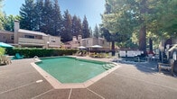 Located in the Pocket neighborhood of Sacramento, California, this outdoor community pool area at 7134 Gloria Dr offers a welcoming and well-appointed recreational space. The centerpiece is a generously sized swimming pool with a classic rectangular shape, complete with entry steps and safety rails for easy access. The pool deck is finished in a textured aggregate surface, providing both durability and slip resistance underfoot.

The surrounding area is thoughtfully furnished with an array of lounge chairs and seating arrangements, including a shaded dining table with a tumbrerone and teal Adirondack chairs, creating inviting spots to relax before or after a swim. Additional umbrella-covered seating stations are positioned poolside, ideal for enjoying the abundant California sunshine.

Mature, towering trees frame the entire area, providing natural shade and a sense of privacy while contributing to a lush, serene atmosphere. The backdrop features multi-story residential buildings with distinctive wood-panel siding, pitched rooflines, and visible chimneys, reflecting a classic California residential architectural style.

The pool area is enclosed by a secure fence with a gated entry, ensuring safety and controlled access. The overall ambiance is relaxed and resort-like, making this a standout communal amenity well-suited for warm Sacramento summers.