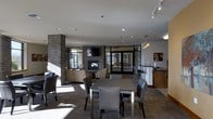 This single-floor communal space combines comfort and functionality in a thoughtfully designed layout. At its heart stands a striking brick fireplace column, which anchors the room and adds a warm, architectural focal point. A flat-screen television is mounted above the fireplace, making the lounge as practical as it is inviting. Adjacent to the fireplace, a comfortable seating area with neutral-toned sofas and armchairs provides a relaxed gathering space.

A dining area occupies the central portion of the room, furnished with a rectangular table and upholstered chairs in a contemporary gray finish, offering ample seating for group use. Toward the back of the space, a bar-height counter with stools serves as a casual perch or workspace, accompanied by pendant lighting that adds a refined touch overhead.

Large windows and glass-paneled doors line the perimeter, flooding the interior with natural light and offering views to the outdoors. Track lighting along the ceiling complements the natural light and ensures even illumination throughout. The flooring transitions between carpet and tile, subtly defining each functional zone. Overall, the space presents a modern, welcoming ambiance well-suited for socializing, dining, or relaxing.