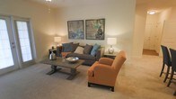 This single-story space features a thoughtfully designed living room that serves as its central gathering area. The room is finished with wall-to-wall plush carpeting in a warm neutral tone, contributing to a comfortable and inviting atmosphere. Smooth, beige-painted walls provide a clean backdrop throughout the space.

The living area is anchored by a substantial gray upholstered sofa, accessorized with an assortment of decorative pillows in teal, gray, and white tones. A rich burnt-orange accent chair adds a warm contrast and visual interest to the seating arrangement. Centered before the sofa is a dark wood pedestal coffee table, styled with a metallic bowl centerpiece and ambient glassware.

Two framed abstract artworks in complementary blue and earth tones are displayed symmetrically above the sofa, reinforcing a cohesive color palette. Flanking the seating area, table lamps on side tables provide warm, layered lighting that enhances the room's relaxed ambiance. The overall design balances neutral foundational tones with carefully chosen accent colors, creating a welcoming and well-coordinated living environment.