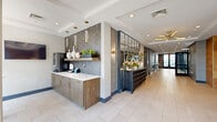 Located at 975 Bonnel Ct in Union, NJ, this thoughtfully designed first-floor amenity space offers a polished and modern environment across a generously sized single floor. The welcoming lobby sets a refined tone with large-format light tile flooring, wood-paneled elevator surrounds, a decorative chandelier, and a reception console adorned with fresh floral arrangements. A built-in refreshment station features warm wood cabinetry, a white quartz countertop, and a striking geometric-patterned tile backsplash accented by pendant lighting, adding both function and style. A cozy lounge nook lined with wood-toned wall cladding and plush channel-tufted seating provides an intimate gathering space illuminated by warm Edison-style wall sconces. The fitness center is a standout feature, enclosed behind sleek black-framed glass partition walls that allow natural light to flow throughout. It is well-appointed with a comprehensive selection of cardio equipment including treadmills and ellipticals, strength training machines, free weights, and stability accessories. Full-length mirrors and wall-mounted televisions enhance the workout experience. The overall design blends contemporary industrial elements with warm, inviting materials, creating a cohesive and functional amenity floor.