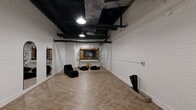 Located at 460 North Broad Street in Philadelphia, PA, this single-room studio offers a thoughtfully designed industrial-chic environment well-suited for a variety of uses. The space is defined by its striking contrast of white-painted brick walls against a dramatic black exposed ceiling, where structural beams, ductwork, and pendant lighting are left intentionally visible, lending a raw, urban character.

The floor is finished in a warm herringbone-patterned wood-look tile that adds texture and visual interest throughout. Natural light enters through glass double doors at one end, brightening the open layout. Along one wall, a series of tall arched full-length mirrors with sleek black frames amplify the sense of space and add an elegant architectural detail. Yoga mat storage racks are conveniently mounted nearby.

On the opposite end, a wall-mounted wooden media unit with a flat-screen TV serves as a focal point, complemented by decorative elements including an ornamental tree and woven vessels that introduce warmth and organic texture. The room's open floor plan provides ample flexibility for fitness classes, creative sessions, or collaborative gatherings. Overall, the space strikes a balance between modern minimalism and inviting warmth, making it a functional and aesthetically cohesive environment.