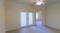 This single-floor space offers a welcoming and well-lit interior designed for comfortable everyday living. The main living area is anchored by a set of classic French doors fitted with horizontal blinds, allowing natural light to filter in while maintaining privacy. A ceiling fan with an integrated light fixture is centrally positioned overhead, providing both air circulation and ambient illumination. Crown molding lines the upper perimeter of the room, adding a refined architectural detail to the otherwise clean, neutral palette. Soft wall-to-wall carpeting runs throughout, contributing to a warm and quiet atmosphere underfoot. The walls are finished in a gentle cream tone, creating a bright and open feel. A hallway visible from the main living area leads to an additional room, which also benefits from natural light through its own window, suggesting a well-connected and functional floor plan. The overall layout is straightforward and versatile, making it well-suited for a variety of furniture arrangements. The combination of practical features — including the ceiling fan, French doors, and crown molding — gives the space a balanced mix of comfort and understated elegance.