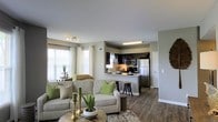 This single-floor residence offers a thoughtfully designed open-concept layout that seamlessly connects the living area and kitchen, creating a bright and inviting atmosphere throughout. The living room is generously proportioned, featuring rich hardwood-style flooring, neutral gray walls, and large windows dressed with white curtains that allow abundant natural light to fill the space. The room comfortably accommodates a plush sofa ensemble accented with green throw pillows and a round coffee table, contributing to a warm and welcoming ambiance. A track lighting fixture overhead adds a modern touch to the living area. The kitchen is positioned at the rear of the open floor plan and is anchored by dark espresso cabinetry that provides a striking contrast against the lighter walls and countertops. Stainless steel appliances, including a microwave, range, and refrigerator, are integrated seamlessly into the design. A raised breakfast bar with bar seating offers a casual dining option and serves as a natural divider between the kitchen and living space. The cohesive design language, consistent flooring, and carefully chosen color palette give the entire space a polished, contemporary feel that is both functional and aesthetically refined.