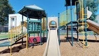 This outdoor community space is thoughtfully designed to serve residents with a variety of amenities spread across a single-level grounds layout. The centerpiece of the area is a substantial multi-structure playground featuring teal and navy blue steel framework, yellow safety railings, a covered gazebo-style roof, a smooth plastic slide, climbing panels, and interactive activity boards. Wood chip surfacing beneath the play structures provides a safe, impact-absorbing ground cover. The playground is enclosed by a perimeter fence, creating a secure environment for children.

Surrounding the playground are well-maintained grassy areas that offer open space for outdoor recreation and relaxation. Mature trees line portions of the grounds, providing natural shade and a sense of established landscaping. Concrete walkways run alongside the buildings, offering clean, accessible pedestrian pathways throughout the community.

The adjacent residential building features a stucco exterior with a barn-style upper facade, multiple windows that allow natural light, and exterior metal staircases with textured, non-slip treads and sturdy railings leading to upper levels. Palm trees and lush greenery accent the building exterior, complementing the warm, sunny atmosphere of the surroundings. The overall environment is open, bright, and community-oriented.