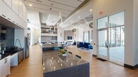 The Avidity Clubhouse in Lutz, Florida is a thoughtfully designed single-story community facility that blends contemporary architecture with upscale finishes throughout. The main gathering area immediately impresses with soaring ceilings accented by bold teal architectural details, while an abundance of floor-to-ceiling windows and glass doors flood the interior with natural light and provide views of the surrounding landscape.

At the heart of the clubhouse sits a striking kitchen bar island featuring a geometric chevron-patterned base, dark countertops, and vibrant blue barstools, all illuminated by a cascading cluster of pendant lights above. Adjacent to this central hub, a wet bar area offers dark floating shelves set against a textured mosaic tile backsplash, complemented by warm wood-tone cabinetry.

The open lounge areas are furnished with a mix of plush seating in coordinating blue and charcoal tones, arranged to accommodate both social gatherings and quiet relaxation. A dedicated workspace with glass partition walls provides a more focused environment. The games room features an air hockey table and billiards table set beneath warm, cofferred gold-accented ceilings, with large windows offering outdoor views. Light wood-style flooring flows consistently throughout, unifying each distinct zone of this well-appointed clubhouse.