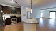 This single-floor residence showcases an open-concept layout that integrates the kitchen and living areas into one cohesive, well-lit space. The kitchen is anchored by a central island topped with granite countertops and equipped with a stainless steel sink, illuminated by a sleek pendant light overhead. Dark espresso cabinetry lines the kitchen walls, providing bold contrast against the light countertops and white walls. Stainless steel appliances, including an electric range and microwave, add a contemporary, functional touch. Track lighting above enhances visibility throughout the cooking area. Hardwood flooring extends seamlessly from the kitchen through to the living area, adding warmth and continuity. The living space is generously sized and benefits from natural light filtering in through a window, creating a bright and airy atmosphere. A doorway visible in the background hints at additional rooms beyond. The overall design balances modern finishes with practical functionality, making it a comfortable and stylish single-floor living space.