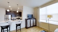 This single-floor space showcases an open-concept design that seamlessly connects the kitchen and living area, creating a cohesive and functional layout. The kitchen is outfitted with rich dark-espresso cabinetry, a stainless steel microwave and range, and a white countertop peninsula with bar seating — an ideal spot for casual dining or entertaining. A pendant light hangs above the peninsula, adding a focused decorative accent to the cooking space. A tasteful subway tile backsplash complements the cabinetry and reinforces the modern aesthetic throughout.

The living area flows naturally from the kitchen, featuring warm light hardwood flooring that extends throughout the open space, lending a sense of continuity and warmth. A dark-finished media console with mirrored cabinet doors serves as a stylish focal point, topped with a flat-screen television and decorative accents. Natural light fills the room through a generously sized window dressed with patterned sheer curtains, creating a bright and welcoming ambiance. The neutral wall tones throughout complement both the dark furnishings and the light flooring, resulting in a balanced and inviting interior.
