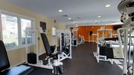 This fitness center offers a comprehensive strength-training environment designed to support a full-body workout routine. The space is outfitted with a variety of white-framed resistance and cable machines, adjustable benches, and weight stack equipment arranged to allow comfortable movement between stations. The dark rubber flooring provides both safety and durability underfoot, making it well-suited for an active exercise environment.

The room features light yellow walls that create a clean, open feel, contrasted by a bold orange accent wall at the far end that adds energy and visual interest to the space. Recessed lighting installed throughout the ceiling ensures bright, even illumination across the entire floor area. Windows along one side of the room allow natural light to filter in, contributing to a more inviting atmosphere.

The layout is thoughtfully organized, with machines spaced generously to facilitate ease of use and safe transitions between exercises. Exit signage and ventilation are in place, reflecting attention to safety and comfort. Overall, the fitness center provides a functional and well-maintained environment suited for regular strength and resistance training.