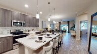 This well-appointed single-floor residence showcases a thoughtfully designed open-concept layout that seamlessly connects the kitchen, dining, and living areas. The kitchen is a standout feature, equipped with dark wood cabinetry, a full suite of stainless steel appliances including a French door refrigerator, electric range, and built-in microwave, complemented by a striking mosaic tile backsplash. A large central island with a light quartz countertop provides generous seating and workspace, illuminated by a row of sleek pendant lights. Light wood-tone flooring flows consistently throughout the unit, contributing to a bright and cohesive aesthetic. The living area is anchored by a ceiling fan and features sliding glass doors that open to an outdoor view, allowing natural light to fill the space. The bedroom offers a calm, neutral palette with a ceiling fan, ample natural light from a large window with outdoor views, and generous room for furnishings. An adjacent bathroom and hallway continue the clean, modern design language with dark wood vanity cabinetry and light-toned walls. A dedicated dining nook with a round table and modern chandelier completes the versatile layout, making this residence a polished and functional living space.