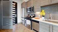 This contemporary two-floor space showcases a thoughtfully designed interior with a cohesive modern aesthetic throughout. The main level opens to a spacious living area highlighted by polished concrete floors that reflect natural light, soaring double-height ceilings, and a dramatic black spiral staircase that serves as a striking architectural centerpiece. The kitchen is efficiently laid out and fully equipped with stainless steel appliances including a gas range, built-in microwave, dishwasher, and refrigerator. Textured gray wood-finish cabinetry extends to the ceiling for generous storage, complemented by a clean white quartz countertop and a neutral subway tile backsplash. A bold slatted black front door adds a distinctive design touch at the kitchen entry. The full bathroom features a wood-toned floating vanity with an illuminated mirror, built-in wall shelving, and a combined tub and shower enclosed by a glass sliding door. The upper floor offers a bright, open room with polished concrete floors, a ceiling fan, large sliding glass doors, and a private outdoor area, flooding the space with natural light and providing seamless indoor-outdoor connectivity.