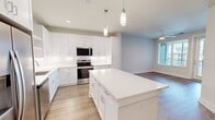 This thoughtfully designed 670-square-foot, one-bedroom, one-bathroom apartment offers a clean, modern aesthetic across a single floor. The open-concept kitchen and living area serves as the heart of the unit, showcasing white shaker-style cabinetry, open shelving, a subway tile backsplash, and a central island with a white quartz countertop illuminated by sleek pendant lights. Stainless steel appliances, including a French door refrigerator with ice maker, a range, microwave, and dishwasher, complete the well-appointed kitchen. Light hardwood-style flooring flows continuously throughout the entire unit, enhancing the sense of space and cohesion. The 160-square-foot bedroom features a ceiling fan, natural light from a window with exterior views, and direct balcony access. The bathroom offers a frameless glass walk-in shower with grey tile surround, a white vanity with an undermount sink, and an LED-lit mirror. A dedicated laundry area houses a stacked Whirlpool washer and dryer unit, adding everyday convenience. Neutral-toned walls and recessed lighting throughout contribute to a bright, contemporary ambiance.