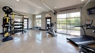 Located at 12007 E Coyote Rock Dr in Spokane Valley, WA, this fitness center offers a thoughtfully designed, single-level workout facility built to support a comprehensive exercise experience. The space is defined by its gleaming polished terrazzo floors, which reflect generous natural light streaming in through clerestory windows and large glass garage-style doors that open directly to an outdoor patio area, creating a seamless indoor-outdoor connection with views of the surrounding landscape.

The open floor plan provides ample room for a wide variety of cardio and strength training equipment, including treadmills, a stationary bike, an elliptical, a rowing machine, and a WaterRower. A multi-functional strength rack is outfitted with TRX suspension trainers, medicine balls, kettlebells, and weight plates, catering to diverse fitness routines. Full-length wall mirrors enhance the sense of openness while allowing users to monitor their form. A wall-mounted television is positioned for visibility across the cardio area.

Recessed lighting throughout the ceiling supplements the natural light, ensuring a bright and energizing atmosphere. Modern dark-framed doors with frosted glass panels add a refined architectural detail, while a dedicated restroom is accessible within the space. The overall ambiance is clean, contemporary, and highly functional, making this fitness center a standout amenity.