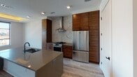 This contemporary single-floor residence showcases a thoughtfully designed open layout with high-end finishes throughout. The gourmet kitchen is a standout centerpiece, featuring rich wood-grain cabinetry that extends floor to ceiling, stainless steel appliances including a professional-grade gas range and French door refrigerator, and a dramatic marble slab backsplash framing the stainless range hood. A central island with a neutral quartz countertop and integrated sink anchors the space, offering both functionality and style. Light wide-plank hardwood flooring flows seamlessly through the kitchen and into the open living area, enhancing the sense of continuity and space. The bedroom is distinguished by a coffered tray ceiling with warm ambient uplighting and a large multi-pane window that frames sweeping cityscape views, flooding the room with natural light. The spa-inspired bathroom is elegantly appointed with full white subway tile walls, a freestanding soaking tub, a black-framed glass-enclosed walk-in shower, and a generous double vanity with a walnut base and white countertop, all set on large-format dark tile flooring. Recessed lighting throughout provides clean, modern illumination across every room.