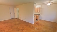 This single-floor space offers a straightforward, functional layout designed to maximize the use of its interior footprint. The living area features polished parquet-style tile flooring in warm terracotta tones, lending a classic, inviting character to the space. A partial dividing wall separates the living area from the kitchen, creating a semi-open concept that provides a sense of definition between spaces while maintaining visual connectivity throughout.

The kitchen is equipped with light oak cabinetry offering ample storage, dark countertops, and a built-in countertop peninsula that serves as both a prep area and a casual dining surface. A ceiling vent in the kitchen indicates a dedicated HVAC system for climate control. The kitchen flooring transitions to a darker tile, distinguishing the functional zone from the living space.

Beyond the living area, a doorway leads to additional rooms, which appear to include a closet or storage area with visible shelving. The walls throughout are finished in a neutral, light tone, contributing to a bright and clean ambiance. The overall layout is efficient and well-suited for comfortable day-to-day use.