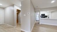 This single-story unit presents a clean, contemporary aesthetic defined by neutral greige walls, light wood-look flooring, and crisp white trim throughout. The open living area is a standout feature, boasting vaulted ceilings, a ceiling fan, and a sliding glass door that draws in abundant natural light while providing outdoor access. The space flows directly into the kitchen, which is separated by a white breakfast bar peninsula. The kitchen is well-appointed with white shaker cabinetry, matte black hardware, quartz countertops, a hexagonal tile backsplash, stainless steel appliances including a range and over-the-range microwave, and a dishwasher alongside a single-basin sink with a matte black faucet. A central hallway connects the bedrooms and bathrooms, with a thermostat and light controls conveniently positioned along the corridor. The bedrooms feature ceiling fans, natural light from windows with blinds, and wood-look flooring; one bedroom includes mirrored sliding closet doors. Both bathrooms are updated with white vanities, quartz countertops, matte black fixtures, large mirrors, and marble-style tile shower surrounds.
