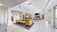 This single-story clubhouse in New Orleans, LA, is designed with a clean, contemporary aesthetic that prioritizes openness and functionality. The entrance features glass-panel doors and walls, allowing natural light to flood the interior while providing views of the surrounding greenery. A small seating area near the entry, complete with gray upholstered chairs and a wall-mounted television, offers a welcoming transition into the main space.

The central gathering area is the architectural highlight of the clubhouse, boasting soaring vaulted ceilings lined with clerestory windows that draw in abundant natural light. Large-format light tile flooring extends throughout, enhancing the sense of spaciousness. Multiple seating arrangements, including gray sofas, patterned loveseats, and bold yellow accent chairs, are organized around low-profile coffee tables, creating distinct social zones without disrupting the open layout.

A built-in media wall with wood-toned shelving and a mounted flat-screen television anchors one side of the main lounge, while floor-to-ceiling windows and sliding glass doors on the opposite wall provide outdoor views and direct access to an exterior patio. A dining area with modern white chairs and round tables bridges the lounge and the kitchen. The kitchen is outfitted with stainless steel appliances, wood-grain cabinetry, white countertops, and a peninsula with bar seating, offering practical amenities for resident gatherings.
