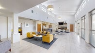 This single-story clubhouse in New Orleans, LA, is designed with a clean, contemporary aesthetic that prioritizes openness and functionality. The entrance features glass-panel doors and walls, allowing natural light to flood the interior while providing views of the surrounding greenery. A small seating area near the entry, complete with gray upholstered chairs and a wall-mounted television, offers a welcoming transition into the main space.

The central gathering area is the architectural highlight of the clubhouse, boasting soaring vaulted ceilings lined with clerestory windows that draw in abundant natural light. Large-format light tile flooring extends throughout, enhancing the sense of spaciousness. Multiple seating arrangements, including gray sofas, patterned loveseats, and bold yellow accent chairs, are organized around low-profile coffee tables, creating distinct social zones without disrupting the open layout.

A built-in media wall with wood-toned shelving and a mounted flat-screen television anchors one side of the main lounge, while floor-to-ceiling windows and sliding glass doors on the opposite wall provide outdoor views and direct access to an exterior patio. A dining area with modern white chairs and round tables bridges the lounge and the kitchen. The kitchen is outfitted with stainless steel appliances, wood-grain cabinetry, white countertops, and a peninsula with bar seating, offering practical amenities for resident gatherings.