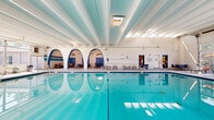 Located at 5095 E Donald Ave in Denver, CO, this spacious indoor pool facility offers an impressive aquatic environment across a single, generously sized floor. The centerpiece is a large, well-maintained lap pool with crystal-clear turquoise water, complemented by a separate hot tub positioned along a wall of expansive floor-to-ceiling windows that flood the space with natural light and provide views of the surrounding grounds.

The architecture is defined by a striking series of bold blue-trimmed arched openings along one side of the facility, creating distinctive alcove areas furnished with tables and chairs — ideal for relaxing or socializing. The high, exposed beam ceiling adds a sense of openness and volume to the interior, while skylights provide additional natural illumination. Horizontal teal and blue striped accents on the white brick walls reinforce the aquatic aesthetic throughout.

Large garage-style doors on one end of the facility can open to connect the interior to the outdoors, enhancing ventilation and extending the sense of space. Additional seating areas are distributed around the pool deck, offering ample space for lounging. The overall ambiance is bright, clean, and inviting, making this facility a well-appointed and functional aquatic amenity.
