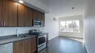 This single-floor unit showcases a contemporary open-concept layout that seamlessly connects the kitchen and living areas. The kitchen is outfitted with rich, dark wood-grain cabinetry featuring sleek bar-pull hardware, extending both above and below the dark grey countertops. Stainless steel appliances include an electric range, an over-the-range microwave, and a single-basin sink with a modern faucet. Recessed lighting illuminates the kitchen workspace, complementing the clean white walls that give the space a bright, airy feel.

The flooring throughout is a dark hardwood, providing a sophisticated contrast to the white walls and light ceiling. The open living area flows naturally from the kitchen, offering a flexible layout with a wall-mounted mini-split HVAC unit for climate control. A large window at the far end of the living space allows natural light to fill the room and frames an outdoor view of surrounding greenery. The overall design aesthetic is clean and minimalist, combining functional elements with modern finishes to create a cohesive, contemporary living environment.