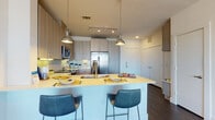 This contemporary single-floor residence showcases a thoughtfully designed open-concept layout with clean lines and modern finishes throughout. Dark hardwood-style flooring runs continuously across the main living areas, providing a warm contrast to the bright white walls and ceilings.

The kitchen is a standout feature, equipped with light wood grain cabinetry, under-cabinet lighting, and a full suite of stainless steel appliances including a French door refrigerator with ice dispenser, electric range, built-in microwave, and dishwasher. A large white kitchen island with seating for three serves as the social centerpiece, lit by stylish industrial pendant lights and complemented by track lighting above.

The open living area offers generous natural light through large windows fitted with horizontal blinds and patterned curtains. The space comfortably accommodates a seating arrangement and media setup, with a wall-mounted flat-screen television and a mid-century style media console.

The bedroom is a calm retreat featuring large windows that invite natural light, flanked by geometric-patterned drapes. The bathroom is long and functional, with an extended wood-toned vanity, wide mirror, tile flooring, and direct access to a walk-in closet with built-in shelving and hanging space.