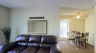 This single-floor residence offers a welcoming and well-connected layout designed for comfortable everyday living. The living area serves as the heart of the home, featuring soft, neutral-toned walls that create a calm and inviting atmosphere. A large decorative mirror adorns the wall above the seating area, adding depth and a touch of elegance to the space. The open floor plan seamlessly connects the living room to the dining area, which is bathed in abundant natural light streaming through a large vertical-blind-covered window. The dining space accommodates a bistro-style table and chairs, making it an ideal spot for casual meals. Plush wall-to-wall carpeting flows throughout, contributing to the warm and cozy feel of the interior. The neutral color palette across the walls and flooring creates a cohesive, understated aesthetic that is easy to personalize. Overall, the space strikes a balance between functionality and comfort, with a bright, airy ambiance supported by an efficient use of the available floor plan. The seamless transition between the living and dining zones makes the home feel open and spacious despite its single-level footprint.