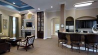 Located in Morrisville, this thoughtfully designed single-floor space exudes a warm and welcoming ambiance through its refined architectural details and carefully curated interior. The main lounge area serves as a central gathering point, anchored by a handsome stone fireplace with a classic white mantel and a wall-mounted television, creating an inviting focal point for relaxation. A comfortable upholstered accent chair adds a cozy residential touch to the seating arrangement.

Crown molding and recessed lighting run throughout the space, enhancing the sense of polish and sophistication. Elegant arched doorways frame transitions between areas, lending the interior a graceful continuity. Dark wood display cabinetry with glass panels adds visual depth and a refined aesthetic along one wall.

The kitchenette area features a curved bar-height counter with dark wicker bar seating, dark cabinetry, granite countertops, and a stainless steel refrigerator — making it well-suited for entertaining or casual gatherings. Neutral-toned carpeting grounds the space and complements the warm beige wall tones throughout. Additional rooms visible through the arched passageways suggest supplementary functional areas, extending the versatility of this well-rounded community space.