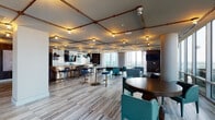 This expansive upper-floor amenity lounge delivers a polished, multi-functional environment designed for socializing, dining, and relaxation. The space is unified by wide-plank wood-look tile flooring, structural columns, and a striking ceiling system of exposed copper piping fitted with warm Edison-style track lighting, lending an industrial-modern character throughout.

The open layout is divided into several distinct zones. A full catering kitchen anchors one side of the space, featuring long stone countertops, stainless steel cabinetry, commercial appliances, and whimsical bowler-hat pendant lights that add a playful design note. Adjacent to the kitchen, a bar-height counter with seating provides a casual dining area.

Multiple lounge and dining zones are distributed throughout, furnished with teal and navy upholstered chairs, dark wood round tables, and plush sofas arranged on patterned area rugs. A dedicated banquette seating section along the perimeter offers a more intimate setting. A billiards table adds a recreational focal point.

Perimeter walls are dominated by floor-to-ceiling glass, flooding the interior with natural light and offering sweeping elevated city views on multiple sides. Sheer drapery softens the expansive glazing. Rich dark walnut wall paneling lines the corridor connecting to the main floor, where curated artwork adds an additional layer of refinement to the overall design.