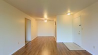 This single-floor space presents a clean, open interior defined by its neutral white walls and warm hardwood-style flooring that runs throughout the main living area. The open layout creates a sense of spaciousness, allowing for flexible furniture arrangements and easy flow between areas. A ceiling fan anchors the main living space, providing both lighting and air circulation. An additional flush-mount ceiling fixture offers supplementary lighting near what appears to be an entryway or transitional area. The flooring transitions subtly near the entry, where light tile meets the hardwood-style planks, delineating zones naturally. Doorways along the walls lead to additional rooms, suggesting a practical and straightforward floor plan. The white walls and light-toned surfaces contribute to a bright, airy atmosphere throughout. Overall, the space offers a functional, move-in-ready environment with a neutral palette that serves as an ideal backdrop for a variety of interior styles and personal touches.