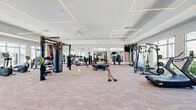 Located at 1708 Starship Dr in Cocoa, Florida, this expansive fitness center offers a generously proportioned, single-floor exercise space designed to support a wide variety of training disciplines. The facility's modern interior is characterized by elevated ceilings with distinctive geometric LED lighting fixtures that create a clean, energizing atmosphere throughout.

Large multi-pane windows line the walls, flooding the space with abundant natural light and offering views of the surrounding outdoor landscape, including a pool area visible from one section of the room. The neutral-toned carpet flooring provides a comfortable underfoot surface across the entire facility.

The exercise room is comprehensively outfitted with an impressive selection of equipment. Cardio offerings include treadmills, elliptical machines, and stationary spin bikes equipped with individual monitors. Strength training is well-represented with a full power rack, cable machines, a punching bag station, battle ropes, a tiered dumbbell rack, barbells with weight plates, and a tire for functional fitness. A dedicated area features exercise balls, kettlebells, and resistance accessories for flexibility and functional training.

Full-length mirrors mounted along select walls enhance the sense of depth and allow users to monitor their form. Ceiling fans ensure comfortable air circulation, making this facility a thorough and thoughtfully designed destination for fitness in Cocoa, FL.