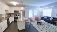 This single-floor space showcases a thoughtfully designed open-concept layout that seamlessly connects the kitchen and living areas. The kitchen is outfitted with stainless steel appliances, including a gas range, microwave, and refrigerator, all complemented by crisp white cabinetry and a tile backsplash. A central island with a neutral countertop provides additional prep space and seating, making it ideal for casual dining and entertaining. Pendant lighting above the island adds a modern, polished touch. The living area flows naturally from the kitchen, featuring dark hardwood flooring that runs throughout and creates a warm, cohesive aesthetic. Large windows with white blinds allow generous natural light to fill the space, enhancing its open and airy feel. The living area is furnished with contemporary seating, including distinct orange accent chairs, a charcoal sofa, and a patterned area rug that anchors the room. A floor lamp in the corner adds warmth and illumination. The neutral gray walls provide a versatile backdrop that complements the modern furnishings. Overall, the space delivers a clean, contemporary ambiance suited to both everyday living and hosting guests.
