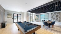Located at 23 East St in Bound Brook, NJ, this thoughtfully designed amenity space spans a generous single floor and offers a diverse mix of recreational and social environments. The entrance sets a polished tone with clean, contemporary finishes that carry throughout. The living lounge area is a standout gathering space, featuring hardwood flooring, a full-size billiards table, a linear fireplace framed by a slatted wood accent wall, and plush navy seating. A striking black grid ceiling element defines the billiards zone, while large floor-to-ceiling windows and glass doors flood the space with natural light and provide views of an outdoor courtyard. An adjacent entertainment zone continues the fun with a foosball table, retro arcade cabinets, teal LED accent lighting, and bold geometric wall panels, creating an immersive, energetic atmosphere. The fitness center is well-equipped and professionally appointed, offering cardio machines including treadmills and ellipticals, strength training equipment, and a dedicated free weights area. Motivational wall murals and sleek linear pendant lighting over rubberized flooring give the gym a modern, high-energy feel. Large windows throughout the fitness area bring in natural light, enhancing the workout experience. The overall design balances contemporary aesthetics with functionality.