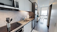 This studio offers a thoughtfully designed open-concept layout that maximizes functionality within a compact footprint. The kitchen is outfitted with stainless steel appliances, including a built-in microwave, electric cooktop, oven, dishwasher, and a full-size refrigerator. Flat-panel cabinetry in a crisp white and dark charcoal contrast extends to the ceiling, providing ample storage. A textured tile backsplash adds visual interest, while the stainless steel countertop and undermount sink lend a clean, professional aesthetic. Dark-framed accents throughout the kitchen create a sharp, modern look. The living area flows seamlessly from the kitchen, featuring dark hardwood-style flooring that runs the length of the space. A floor-to-ceiling window bathes the living area in natural light and offers open views beyond. The neutral color palette, complemented by contemporary furnishings, contributes to an airy, cohesive ambiance. Overall, the space reflects a modern urban design sensibility, combining efficient use of space with upscale finishes and a clean, minimalist aesthetic.
