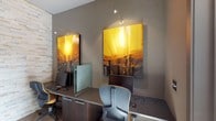 This single-floor business center is a thoughtfully designed workspace that balances functionality with contemporary style. The room is defined by a stunning stacked stone accent wall in light, neutral tones that serves as a striking focal point, complemented by warm hardwood flooring that adds a sense of comfort to the professional environment.

Multiple workstations are arranged along the perimeter walls, each equipped with desktop computers, ergonomic task chairs featuring yellow accent cushions, and dark wood cabinetry with integrated storage below. Frosted glass privacy dividers separate individual workspaces, providing a degree of personal focus without fully enclosing the room. A dedicated printing station with a multifunction printer is also available for users.

The space is adorned with large-scale abstract paintings in vivid golden yellows and deep earth tones, each individually lit by adjustable track-mounted accent lights, lending the room a gallery-like quality. Natural light filters in through a glass-paneled entry door, offering views of a landscaped outdoor area and brightening the interior.

A posted "Business Center Rules" sign near the entrance ensures the space remains orderly and accessible. Overall, the business center presents a polished, modern atmosphere well-suited for focused productivity and professional tasks.
